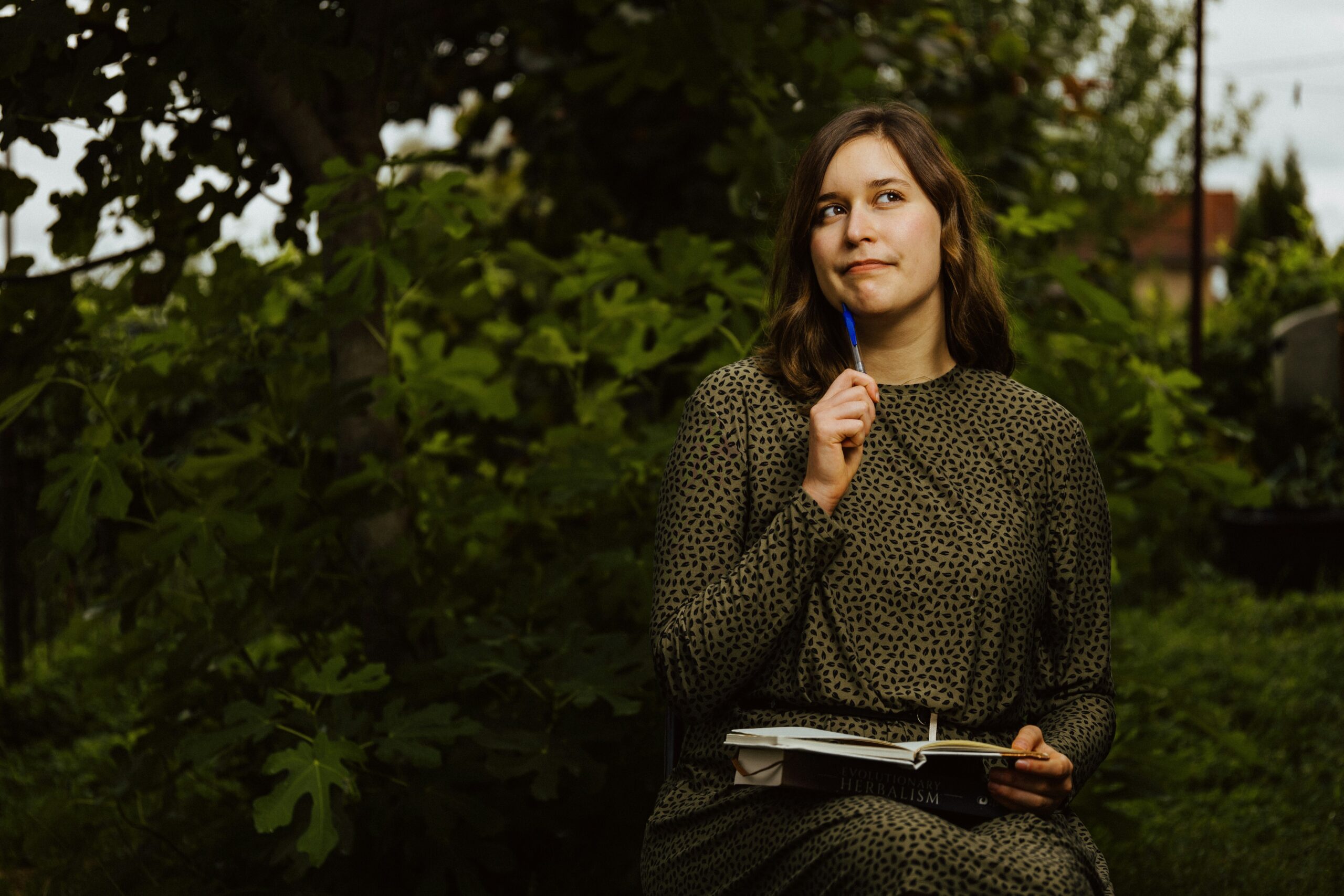Frau mit Notizbuch auf dem Schoß im Halbprofil, hält einen Stift ans Kinn und schaut nachdenklich nach oben, im Hintergrund Wald.